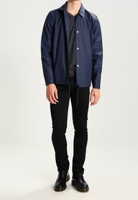 Granatowa wodoodporna kurtka z zapięciem na zatrzaski, lekki materiał i prosty krój. Połączona z czarnymi spodniami i czarnymi sznurowanymi butami.