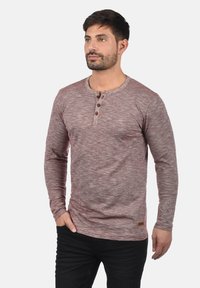 Hosszú ujjú henley póló bordó foltos anyagból. Kerek nyakkivágással, három gombos részlettel és egy apró márkacímkével a szegélyen.