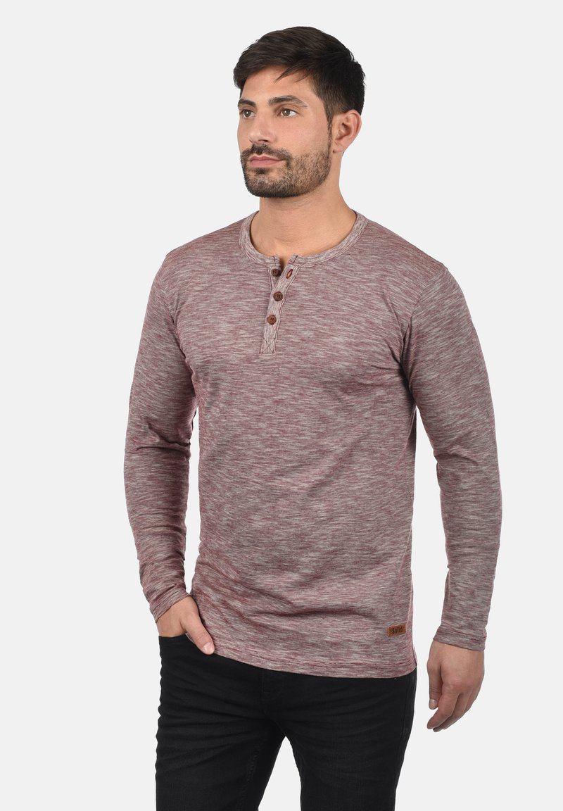 Langermet henley-skjorte i et bordeaux-kornet stoff. Har rund hals, tre knappdetaljer og en liten merkeetikett på kanten.