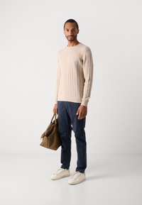 Άνδρας ντυμένος με beidge ζακέτα με πλέξη καλωδίου, ναυτικές παντελόνες, λευκά αθλητικά παπούτσια, κρατώντας καφέ δερμάτινη τσάντα ταξιδιού, στέκεται απέναντι σε λευκό φόντο.