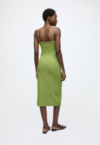 Zaļu teksturētu audumu vidēja garuma kleita ar plānām lencītēm un pieguļošu siluetu, ar taisnu kakla izgriezumu, kas sniedzas līdz pusstilbam, pasniegta kopā ar melniem plakanajiem apaviem.