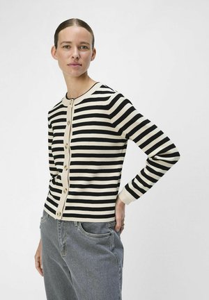 Gilet noir et crème rayé avec des manches longues, un col rond et une fermeture à bouton sur le devant. Porté sur un jean gris, avec une texture tricot lisse.