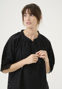 Juoda tunika su gėlių siuvinėjimu, raukšlėtu kaklu ir kutais. Laisvas siluetas su išpjovomis ant rankovių ir apatinės dalies.