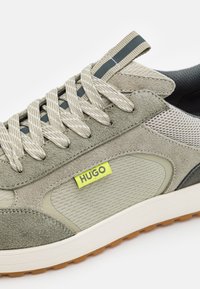 Grågröna sneakers med en blandning av mocka och nätmaterial, randiga snören och en framträdande gul etikett med texten "HUGO." Gummisula.