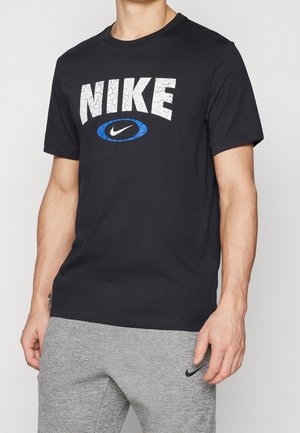 Person trägt ein schwarzes Nike-T-Shirt mit rissigem weißem Logo und blauem Oval, kombiniert mit grauen Jogginghosen mit kleinem schwarzem Nike-Swoosh.