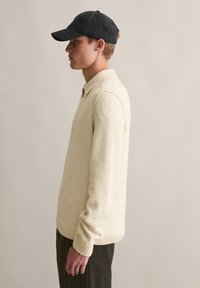 Sweter dzianinowy w jasnym kremowym kolorze o teksturowanym wykończeniu, z zapięciem na guziki przy kołnierzu oraz ściągaczami na rękawach, połączony z ciemnozielonymi spodniami z korduroy.