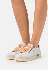 Baskets en daim grises clairs avec des lacets oranges, fermeture éclair sur le côté et accent logo vert. Comprend une semelle texturée et un design bas.