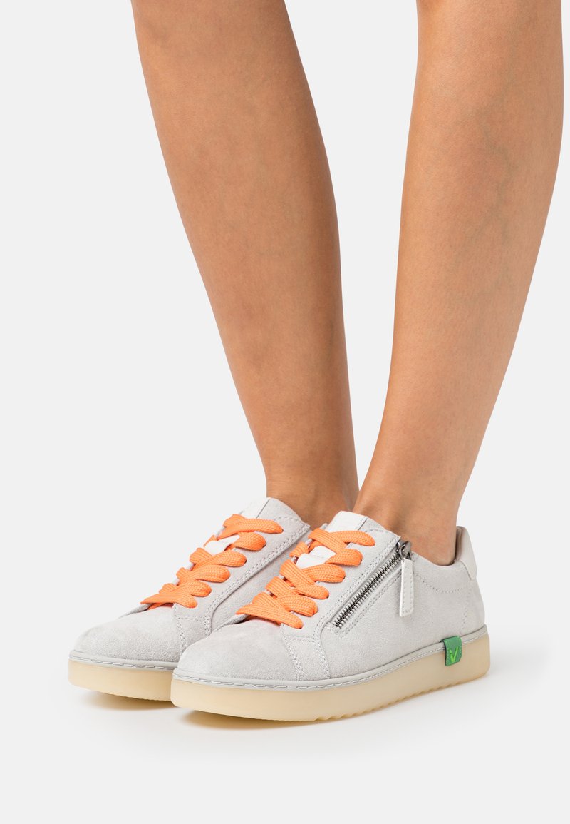 Baskets en daim grises clairs avec des lacets oranges, fermeture éclair sur le côté et accent logo vert. Comprend une semelle texturée et un design bas.