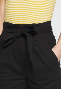 Černé kalhoty s vysokým pasem a uvázaným páskem. Mají přední kapsy a hladký povrch. Osoba má na sobě tílko se žlutými a bílými pruhy.