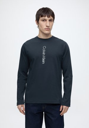 Mladý muž nosící tmavé tričko s dlouhým rukávem Calvin Klein s vertikálním logem, stojí před jednobarevným bílým pozadím.