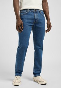 Pantalones vaqueros de mezclilla azul con corte recto, cinco bolsillos y detalles de costura sutiles. Combinados con zapatillas en tono blanco roto. La textura parece suave.