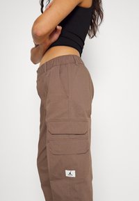 Bruine cargobroek met een gestructureerd oppervlak, voorzien van een elastische tailleband en twee zijzakken. Inclusief een logopatch nabij de rechterzak.