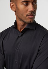 Zwarte lange mouwen shirt van gladde stof, met een puntige kraag, zes knopen aan de voorkant en een getailleerd ontwerp.