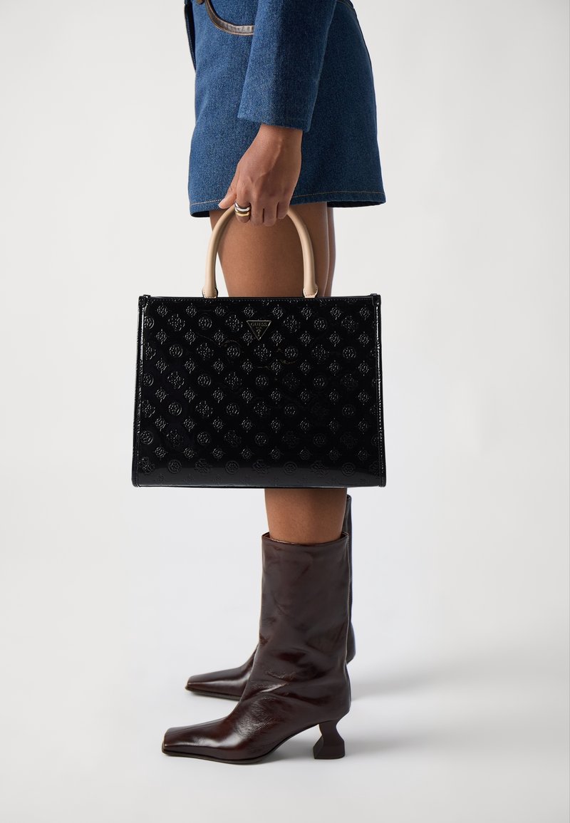 Personne portant une robe bleue et des bottes marron tenant un sac à main noir à motifs avec des poignées beiges sur un fond uni.