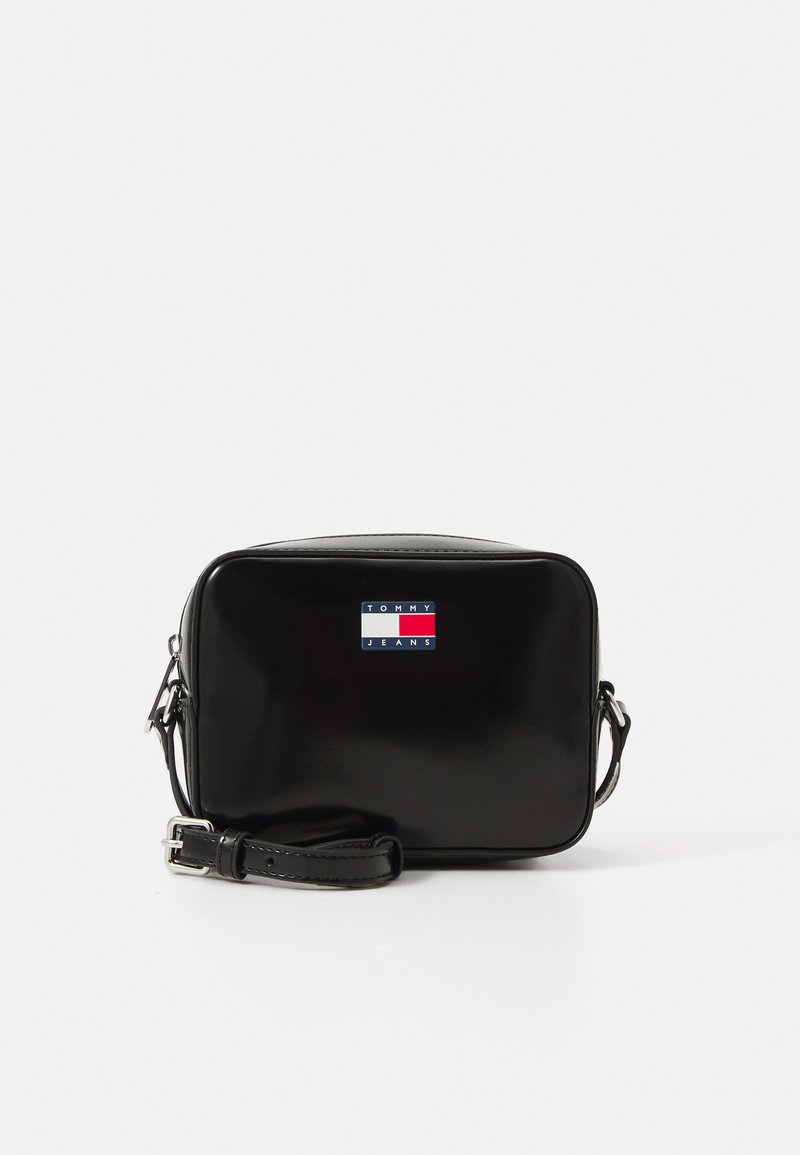 Bolso cruzado de charol negro con forma rectangular, que presenta un parche de logo en bloque de colores con acentos rojos, blancos y azules.