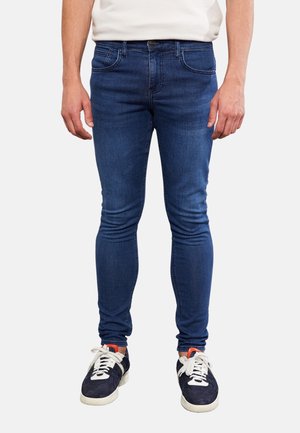 Mężczyzna ubrany w dopasowane ciemnoniebieskie dżinsy i czarne trampki z białymi sznurówkami, stojący na jednolitym tle.