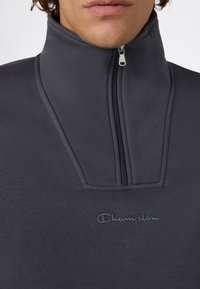 Primo piano di una persona che indossa un pullover Champion grigio scuro con colletto alto zip e logo ricamato sul petto.