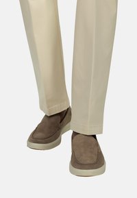 Pantalon beige porté avec des chaussures en daim marron à enfiler, présentant des semelles en caoutchouc blanc, sur un fond blanc uni.