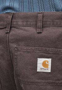 Καφέ τσέπη παντελονιού Carhartt με ορατό λογότυπο της μάρκας, συνδυασμένη με μπλε υφασμάτινο πλεκτό τοπ.