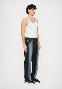 Moške kavbojke z ravnimi hlačnicami v temno modrem denimu z izpranim videzom, kombinirane z belo majico brez rokavov in črnimi športnimi copati.