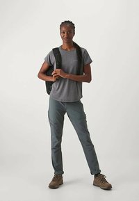 Pelēks īsām piedurknēm krekls, tumši zaļas bikses un brūni pārgājienu zābaki. Melna mugursoma ar regulējamām siksnām. Stāv uz neitrāla fona.