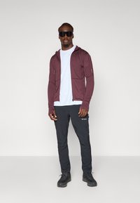 Maronfarvet zip-up hættetrøje over en hvid t-shirt, parret med sorte bukser med sidelommer. Sorte sko fuldender outfittet.