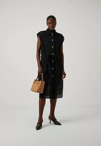 Γυναίκα που φοράει ένα αμάνικο μαύρο φορεματάκι midi με κουμπιά και ζώνη, κρατώντας μία καφέ τσάντα και με μαύρα χαμηλοτάκουνα παπούτσια.