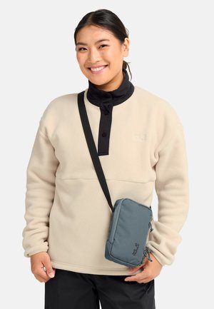 Jonge vrouw glimlachend, gekleed in een beige fleece met een zwarte kraag, zwarte broek en een kleine grijze crossbodytas met een zwarte band.