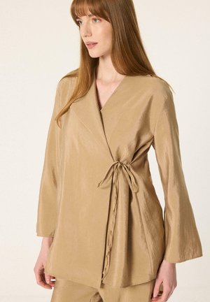 Femme portant un chemisier cache-cœur beige à manches longues avec un nœud sur le côté, debout devant un fond clair et uni, regardant sur le côté.