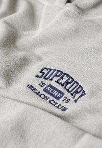 Felpa grigia con testo ricamato blu navy che legge "SUPERDRY 19 SURF 79 BEACH CLUB". Realizzata in tessuto morbido con una texture liscia.