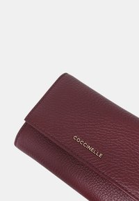 Portafoglio in pelle bordeaux con superficie texture. Dotato di chiusura a pattina magnetica e logo del marchio impresso in oro. Forma rettangolare.