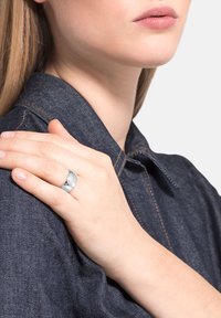 Anillo de plata con una superficie texturizada y pulida, llevado en una mano reposando sobre una tela de mezclilla con costuras visibles y de color azul oscuro.