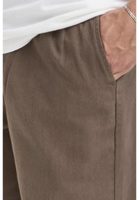 Pantaloni di cotone marroni con una texture liscia, tasca laterale e una cintura elastica parzialmente visibile. Cuciture rinforzate lungo le cuciture.