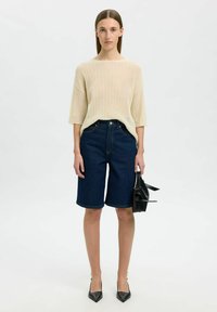 Beige Rippstrickpullover mit Dreiviertelärmeln, kombiniert mit dunkelblauen Denim-Bermudas. Das Model hält eine schwarze Handtasche und trägt schwarze High Heels.