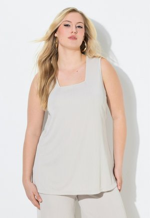 Blonde Frau, die ein ärmelloses, hellbeiges Oberteil und passende Hosen trägt, posiert vor einer weißen Wand mit großen Creolen.