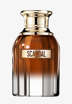 Bottle of Scandal perfume with amber-colored liquid and shiny gold cap, label centered with bold brown and copper text.
