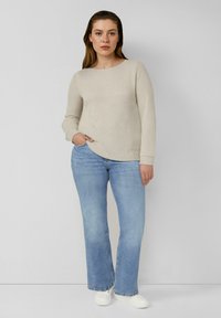 Jasnobeżowy teksturowany sweter z długimi rękawami, w połączeniu z jasnoniebieskimi rozszerzanymi dżinsami i białymi trampkami. Model stoi z ręką w kieszeni.