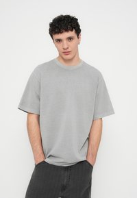 Young man with short curly hair wearing a loose gray t-shirt and dark jeans, hands in pockets, standing against a plain white background.