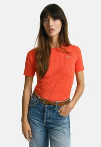 Orange kabelstrikket kortærmet sweater med rund halsudskæring, båret med blå højtaljede jeans og et brunt bælte. Enkel broderet logo detaljer.