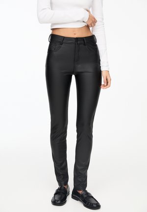 Zwarte kunstleren broek met een slim fit, voorzien van een knoopsluiting, ceintuurlussen en een soepele textuur. G gedragen met een witte top met lange mouwen.