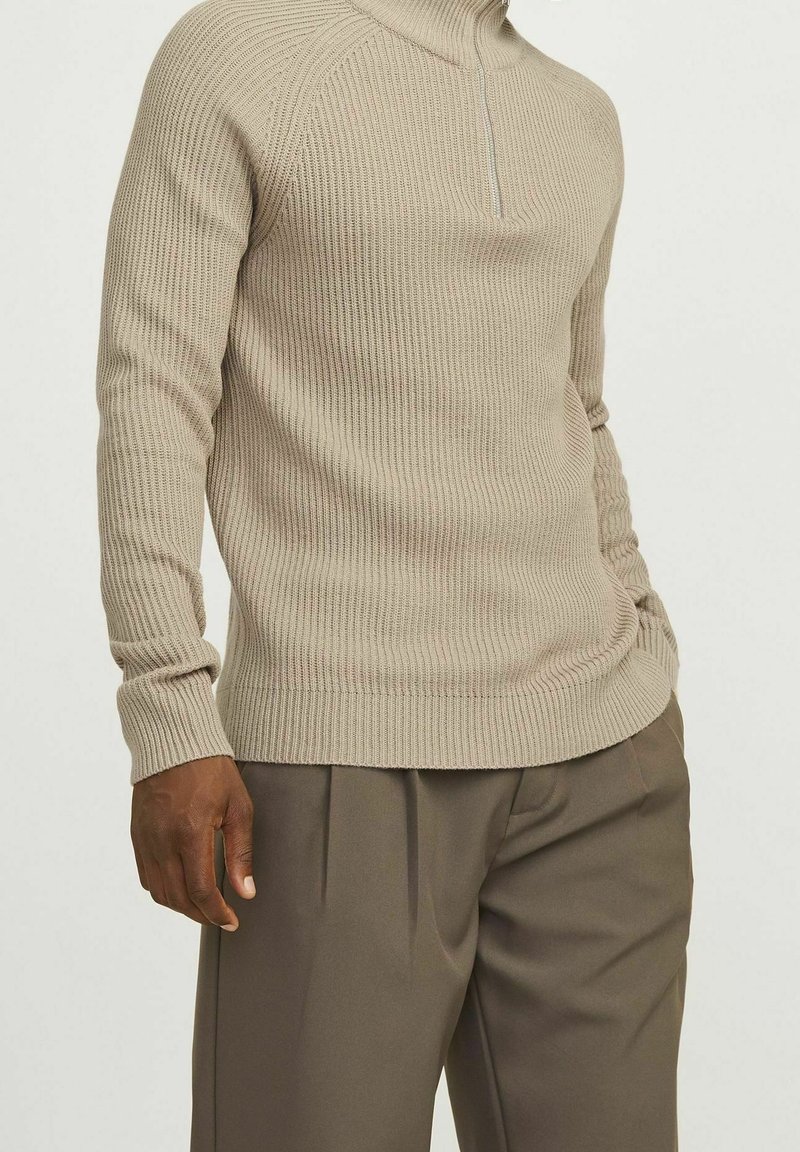 Maglione beige a coste con collo alto e zip a un quarto, abbinato a pantaloni scuri marroni su misura. Il maglione presenta maniche lunghe e tessuto texturizzato.