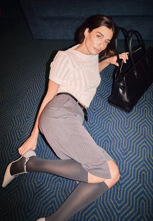 Bolso de mano negro, blusa de punto rosa claro, culottes a rayas grises, medias largas grises y tacones beige sobre un fondo azul con patrones.