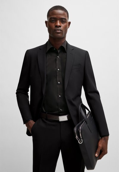 Un costume noir sur mesure avec une chemise noire à boutons, une ceinture marron et une mallette noire texturée. Tissu lisse et éléments de design élégants.