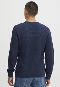 Marineblau gestrickter Pullover mit strukturierten Mustern, langen Ärmeln sowie gerippten Bündchen und Saum. Getragen mit hellblauen Jeans.