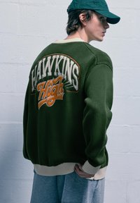 Maglione a maglia verde con la scritta "HAWKINS High" ricamata in bianco e arancione. Presenta un colletto e un orlo a costine color crema.