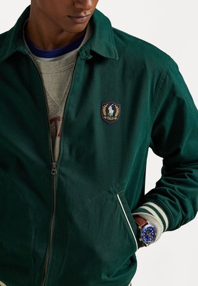 Veste verte en coton avec fermeture éclair à l'avant, ornée d'un patch logo et de poignets rayés. Portée par-dessus un pull gris, complétée par une montre bleue.