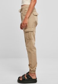 Beżowe spodnie bojówki z elastycznymi mankietami i ściągaczem w pasie, wyposażone w wiele bocznych kieszeni oraz o luźnym kroju. Noszone z czarnymi sandałami.