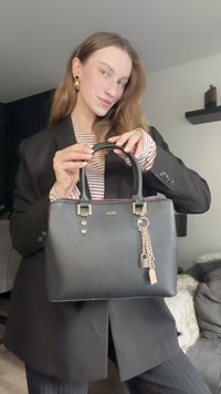 Juoda sintetinė rankinė su dviem viršutinėmis rankenomis, auksinės spalvos detalėmis, trimis akcentinėmis smeigtukėmis ir raktų pakabuku su mažo užrakto detale.