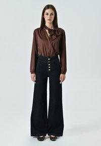 Blusa marrone con dettagli a fiocco, abbinata a jeans in denim scuro a vita alta e gamba larga, con bottoni e cuciture dorate.