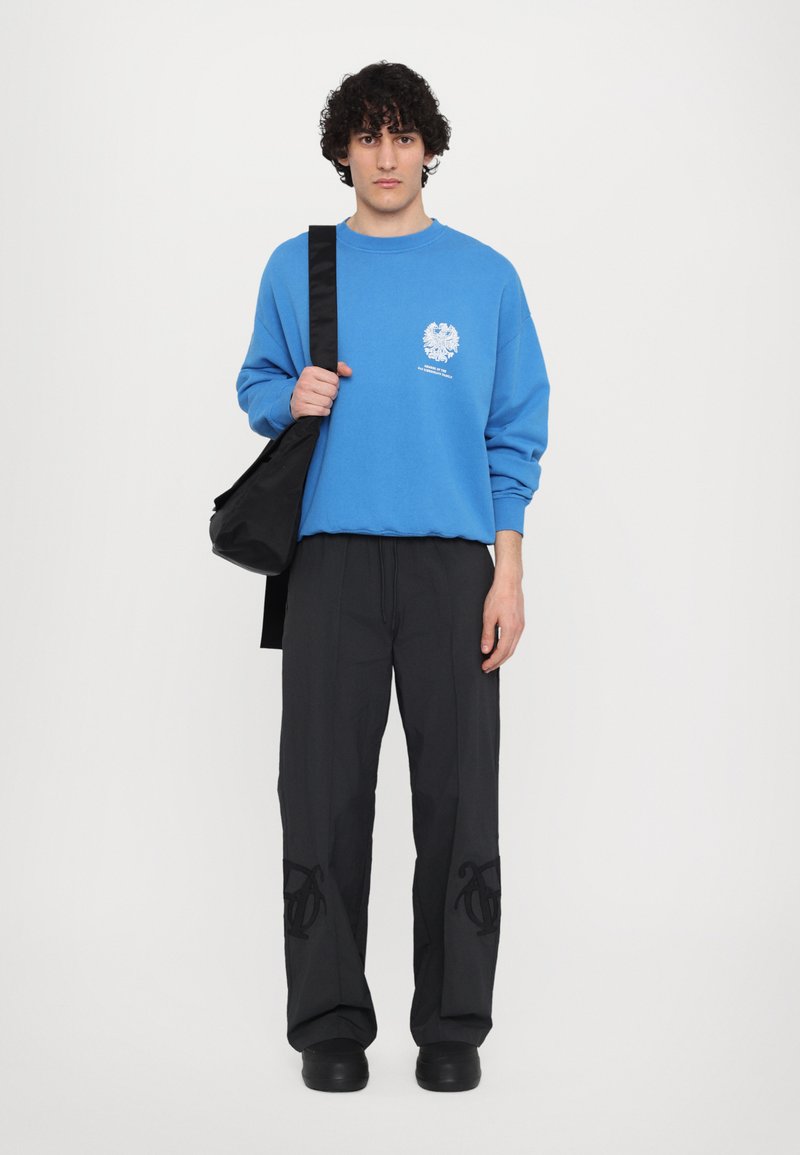Blaues Sweatshirt mit einem kleinen weißen Grafik, kombiniert mit lockeren schwarzen Hosen mit dunkler Stickerei und schwarzen Schuhen. Enthält eine schwarze Tasche.